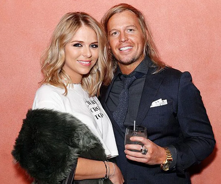 A couple smiling and posing in front of a peach-colored wall; one holds a drink, the other wears a fur coat.