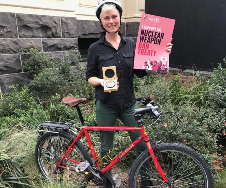 "Person holding 'Nuclear Weapon Ban Treaty' sign and medal, standing beside a red bicycle, wearing a helmet."