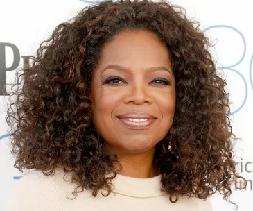 A person with curly hair smiling, wearing a light sweater in front of a light background with text.