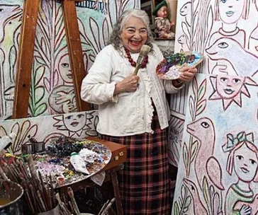 An artist smiles, holding a paintbrush and palette, surrounded by colorful, child-like paintings of faces and animals.