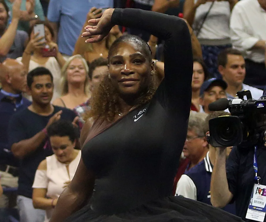 Serena Williams on court at the US Open in a black outfit, surrounded by a cheering crowd and cameras.