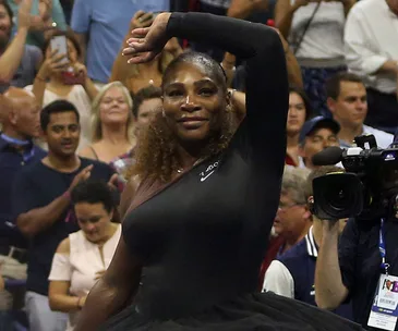 Serena Williams on court at the US Open in a black outfit, surrounded by a cheering crowd and cameras.