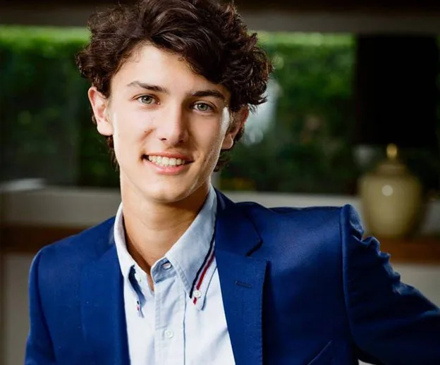 A young person in a blue blazer smiles warmly, standing indoors with a blurred background.