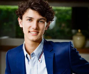 A young person in a blue blazer smiles warmly, standing indoors with a blurred background.