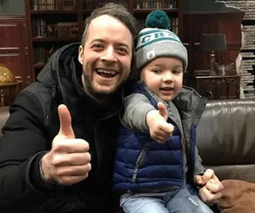 "Smiling man and child with thumbs up, sitting on a brown leather couch in a cozy library setting."