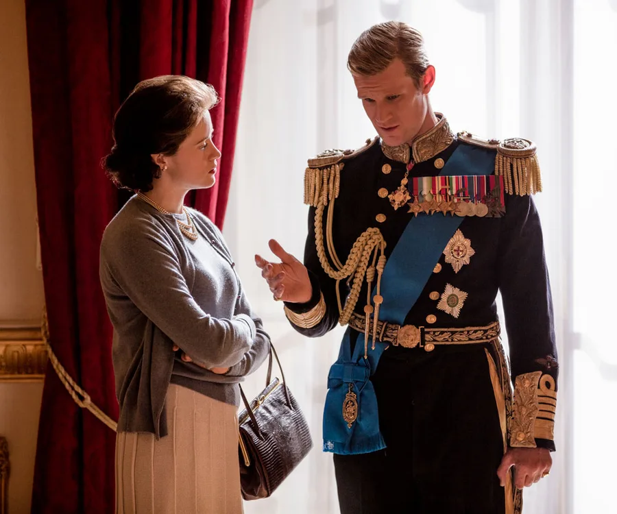 A woman and a man in military uniform converse, set against rich red curtains, in a scene from "The Crown."