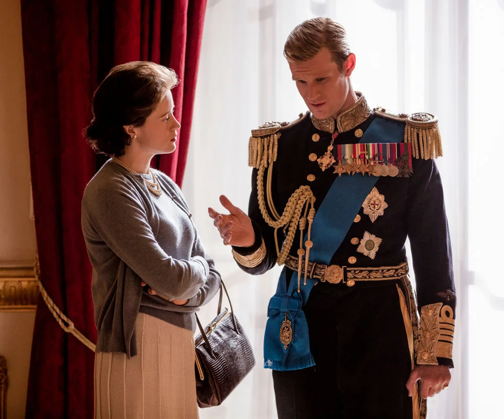 A woman and a man in military uniform converse, set against rich red curtains, in a scene from "The Crown."