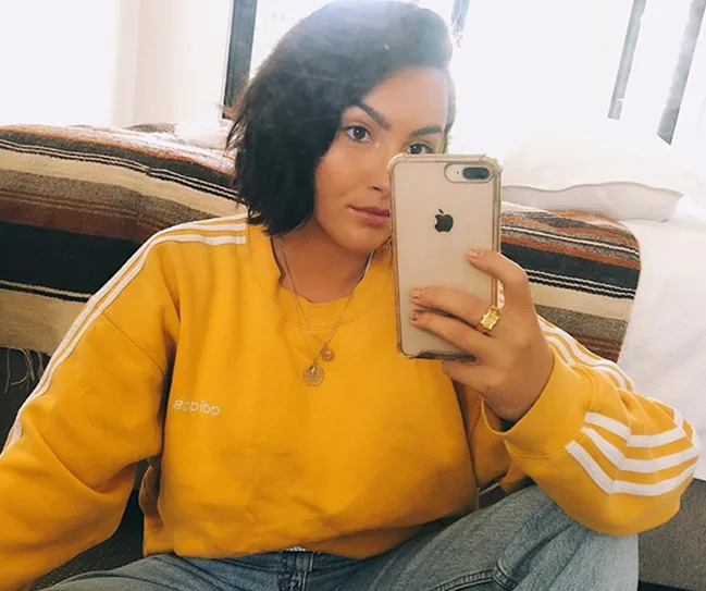 Person taking a mirror selfie wearing a yellow Adidas sweatshirt, gold necklaces, and holding an iPhone.