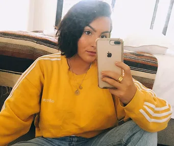 Person taking a mirror selfie wearing a yellow Adidas sweatshirt, gold necklaces, and holding an iPhone.