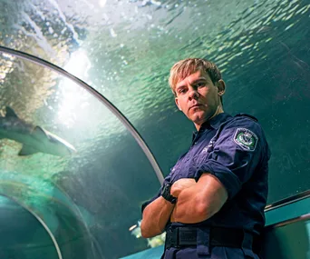 Officer in uniform inside an aquarium tunnel with a shark swimming above.