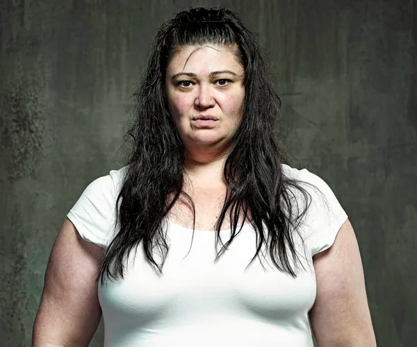 A woman with long dark hair in a white shirt stands against a plain gray background, looking forward.