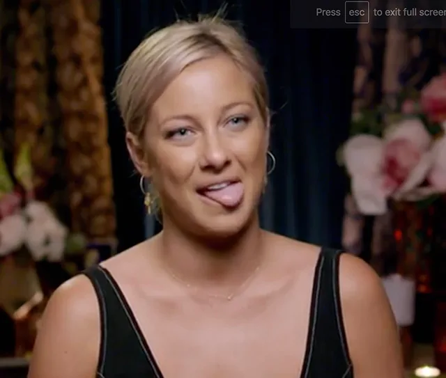 Woman with blonde hair and black top sticking out her tongue, in a room with floral decor.