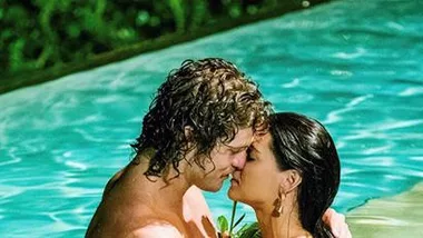 A man and woman share a romantic moment, standing close in a swimming pool with lush greenery in the background.