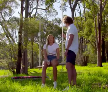 Man in white shirt speaks to a woman on a swing in a sunlit forest clearing.
