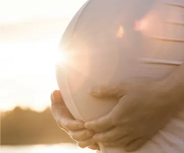 Pregnant belly outdoors with sunlight, hands gently holding stomach.