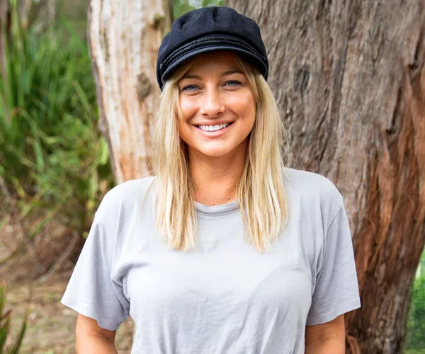 A woman with blonde hair wearing a black cap and gray shirt stands smiling in front of a tree.