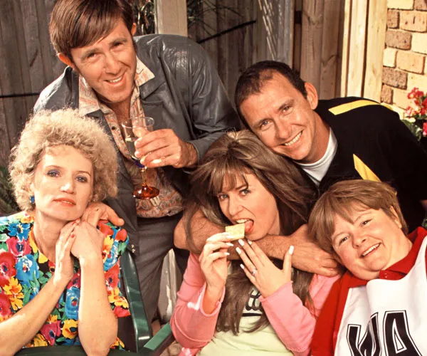 Five people sit together, smiling. One woman eats a cracker. They appear in a casual, colorful setting, suggesting a lively social scene.