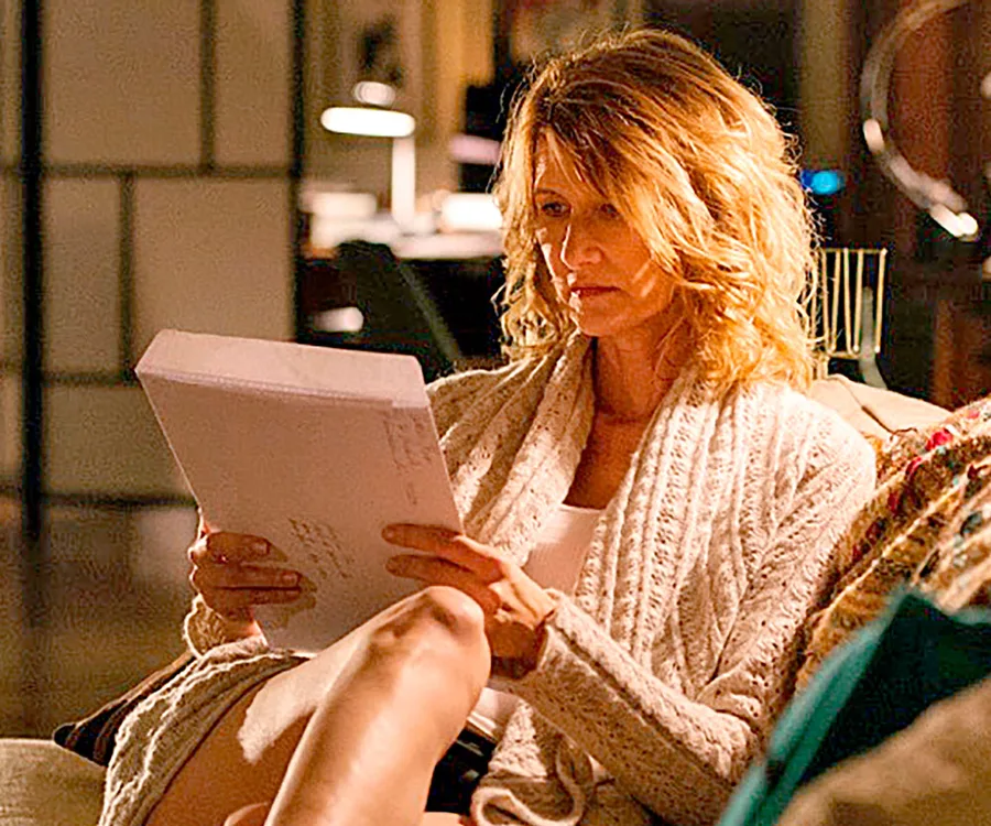 A person with wavy hair sits on a couch, reading a script intently, wearing a cozy sweater in a softly lit room.