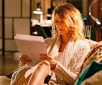 A person with wavy hair sits on a couch, reading a script intently, wearing a cozy sweater in a softly lit room.