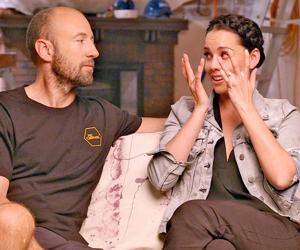 Man and woman sit on a couch; woman is emotional, wiping tears, while man looks on with concern.