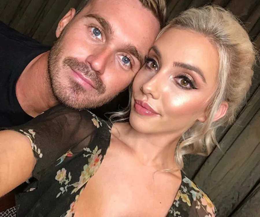 Two individuals posing for a selfie, one wearing a floral outfit, both with smiles, standing closely together.