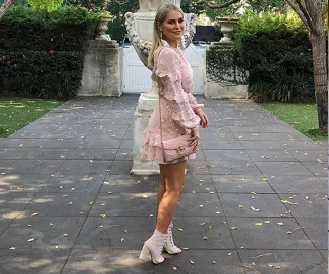 Woman in pink frilly dress and ankle boots poses outdoors, holding a beige purse, with greenery and stone sculptures in background.