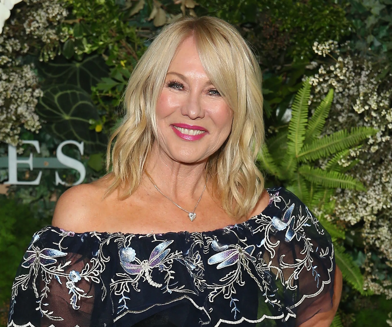 Blonde woman smiling, wearing a floral off-shoulder dress, standing in front of a leafy green background.