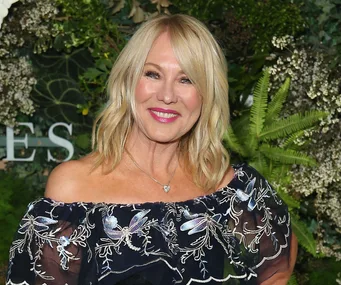 Blonde woman smiling, wearing a floral off-shoulder dress, standing in front of a leafy green background.