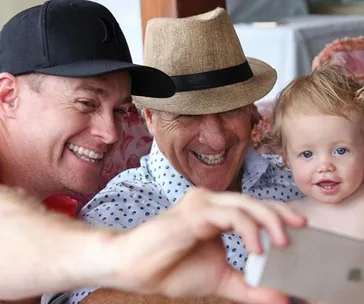 Group selfie with a man in a cap, an older man in a fedora, and a smiling toddler.