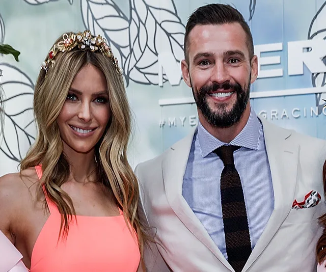 Two people smiling at an event, one wearing a coral outfit with a floral headpiece, the other in a light suit and tie.