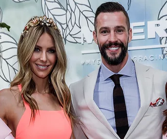 Two people smiling at an event, one wearing a coral outfit with a floral headpiece, the other in a light suit and tie.