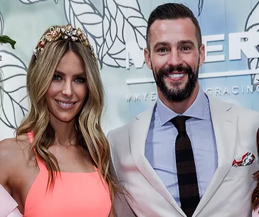 Two people smiling at an event, one wearing a coral outfit with a floral headpiece, the other in a light suit and tie.