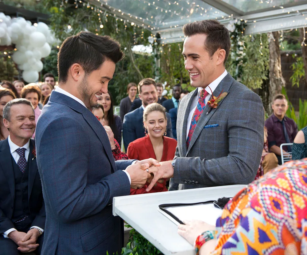 Same-sex wedding scene from "Neighbours," with guests smiling as two men exchange rings under a decorated canopy.