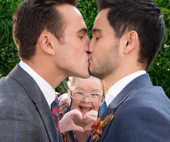 Same-sex wedding scene with a joyful guest making a heart symbol in the background.
