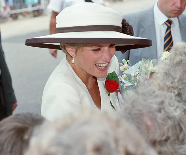 Princess Diana smiling in a wide-brimmed hat, holding a bouquet of flowers, interacting with a crowd.