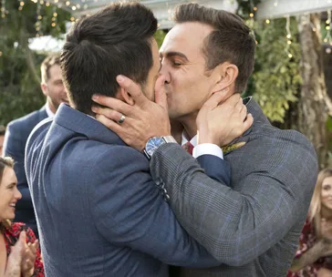 A same-sex wedding scene from "Neighbours" with two grooms in suits sharing a kiss, surrounded by cheering guests.