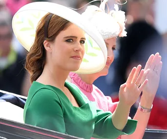 A woman in a green outfit and wide-brim hat waves from a carriage during a formal event.