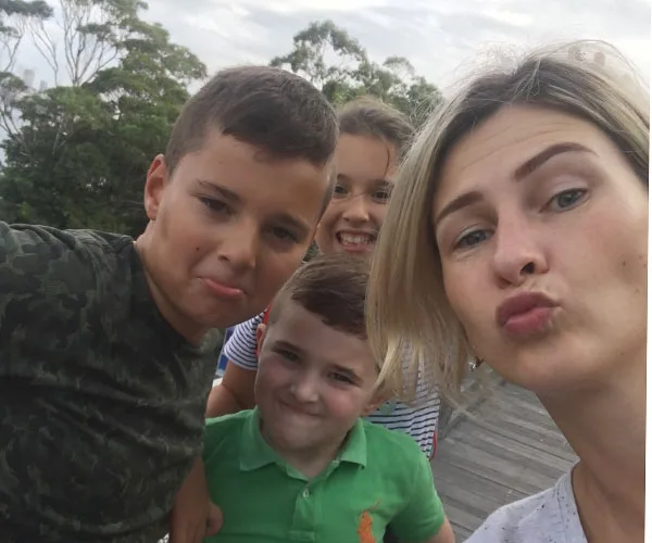 Group selfie with a woman and three children making playful faces outdoors.