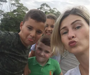 Group selfie with a woman and three children making playful faces outdoors.