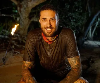 Man with tattoos smiling, seated outdoors in a wet shirt, tribal necklace, and torch lit behind, on "Australian Survivor."