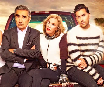 "Three characters from Schitt's Creek sitting on a bench in front of a truck, with sunset in the background."