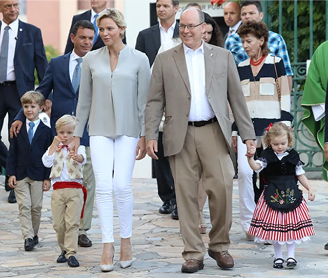 Monaco's royal family, including twins Gabriella and Jacques, walk together outdoors, attended by officials and family.