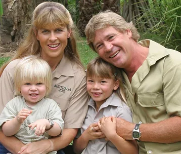 A family smiles outdoors, wearing zoo uniforms, surrounded by greenery.