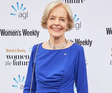 A smiling woman in a blue dress stands in front of a "Women's Weekly" backdrop at an awards event.