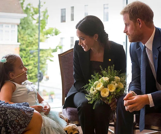 Prince Harry and Meghan Markle smiling and talking with a young girl, Meghan holding a bouquet of flowers.