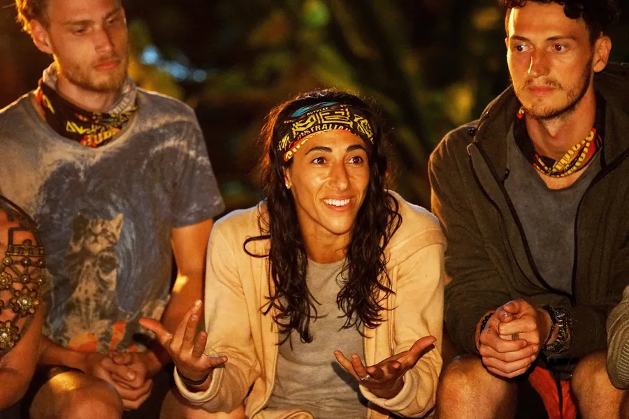 "Person with Australian Survivor headband speaking, seated between two others at night, expressing with hands."
