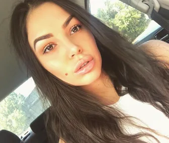 A woman with long brown hair takes a selfie in a car, wearing a white top.