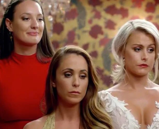 Three women stand together in a formal setting, displaying various emotional expressions.