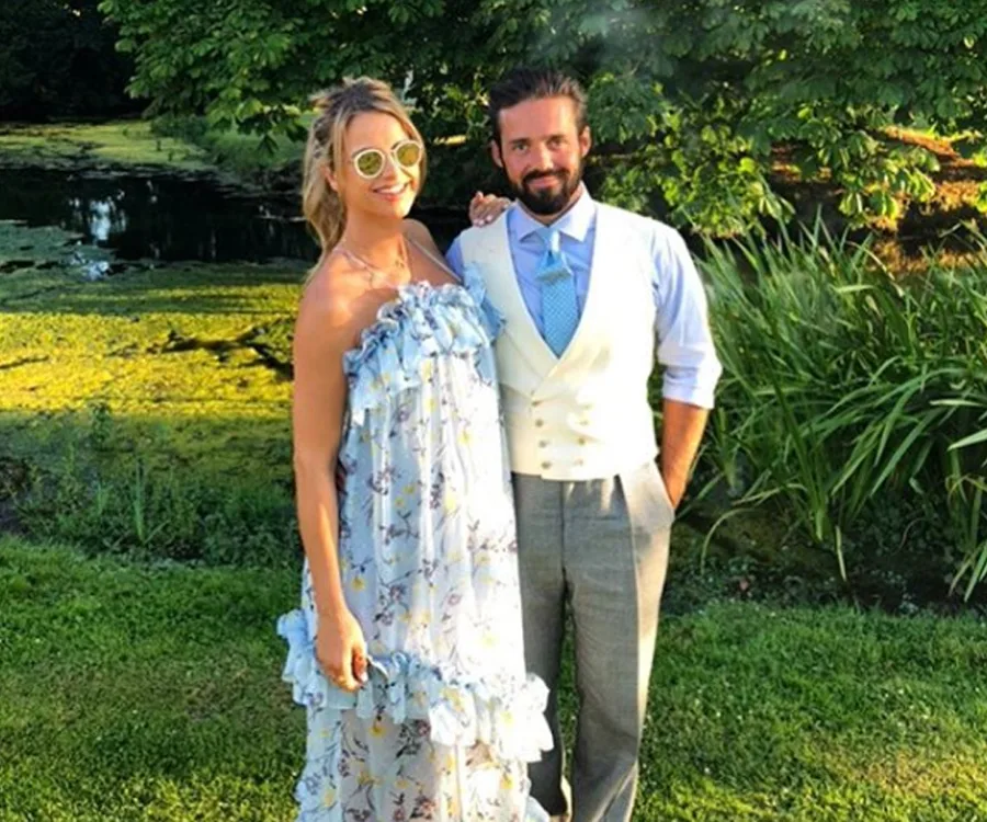 A couple poses in a garden; the woman wears a floral dress and sunglasses, the man wears a suit with a vest and tie.