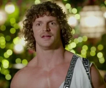Man in Greek-style toga with curly hair and mustache against a bokeh light background.
